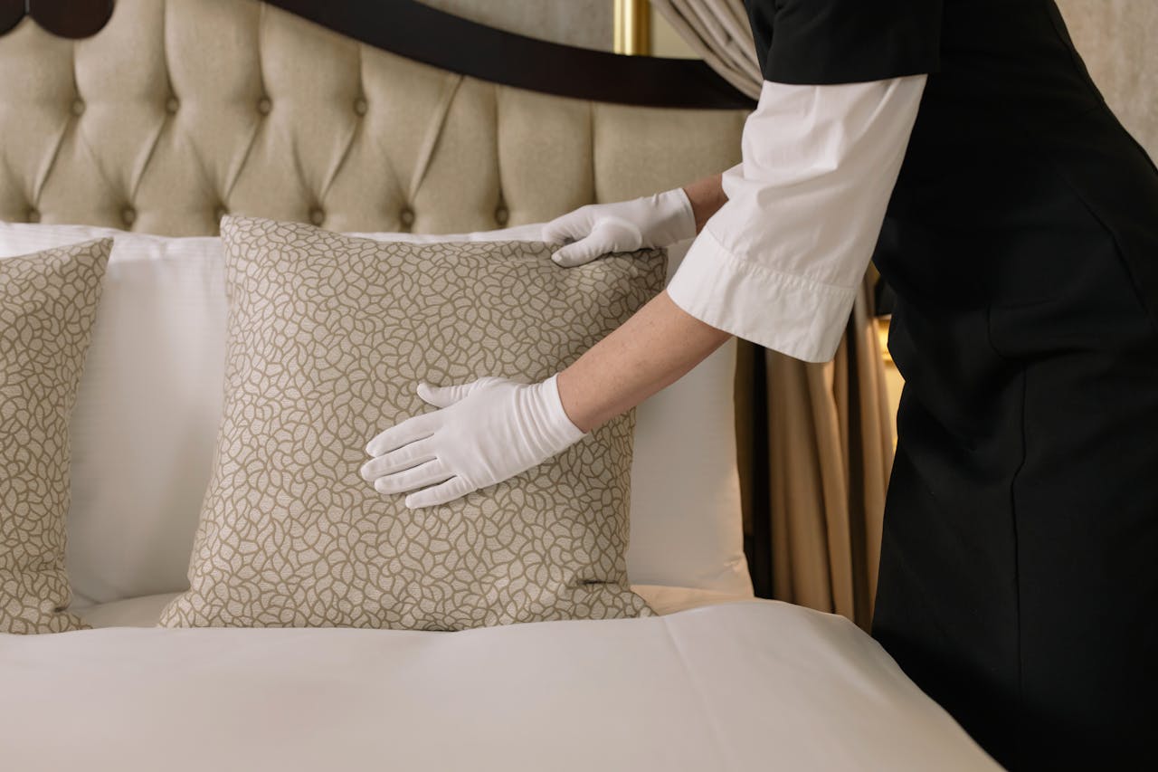 A housekeeper wearing gloves arranges pillows on a luxurious bed in a hotel room.